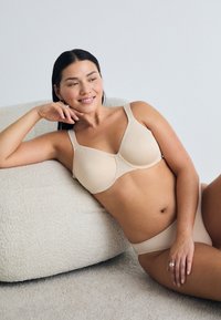 Nainen makaa teksturoidulla beige-sohvalla päällään matching beige rintsikat ja alushousut, hymyillen ja käsi poskellaan.