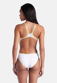 Vit bikini med guldaccenter, med en racerback-top och högmidjade bottnar. Slät textur, minimalistisk design, inga uppenbara mönster.
