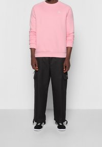 Vaaleanpunainen sweatshirt, jossa on ribbatut hihansuut ja helma, yhdistettynä mustiin cargo-housuihin, joissa on sivutaskuja, ja mustiin lenkkareihin, joissa on valkoiset raidat.