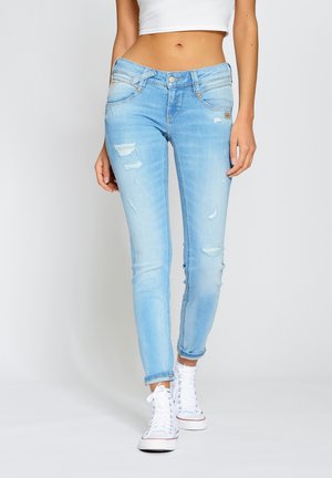 Jeans slim fit bleu clair déchirés portés avec des revers roulés et des baskets montantes blanches, modèle montré de la taille aux pieds.