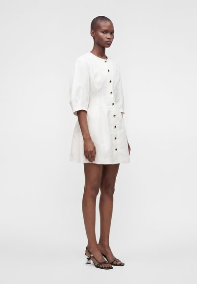 Femme portant une robe courte blanche boutonnée avec des manches bouffantes et des sandales à talons ouvertes noires, debout devant un fond clair uni.
