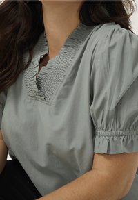 Vrouw in een lichtgrijze blouse met een gerimpelde V-hals en kort met elastische manchetten. Afgebeeld van schouders tot taille.