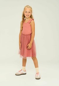 Rosa linne med volangärmar, lager av tylkjol och vita sneakers med rosa ränder. Mot en enkel ljus bakgrund.