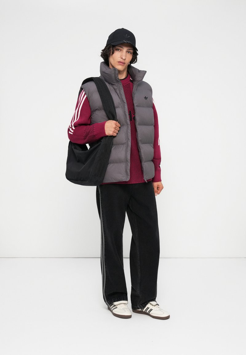 Joven vestido con un chaleco acolchado gris, suéter burdeos, gorra negra, pantalones negros con rayas, zapatillas blancas, sosteniendo un bolso negro de hombro.