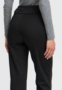 Black high-waisted trousers with a smooth texture, featuring a wide waistband and straight leg design, seen from the back.
