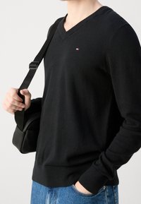 Musta v-kaula-aukkoinen neule pehmeästä kankaasta, jossa pieni logo rinnassa. Yhdistetty sinisiin farkkuihin ja mustaan olkalaukkuun.