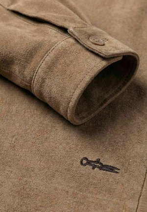 Brun semsket jakkeerm med knappet mansjett og en liten brodert svart dolklogo på stoffet nær mansjetten.