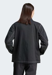 Osoba s rameny dlouhými tmavými vlasy, oblečená v černém kabátě s pruhy a odpovídajících kalhotách, stojící zády s rukama v kapsách.