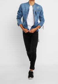 Person som har på sig en blå denimjacka över vit skjorta, svarta tajta jeans och svarta sneakers, som stänger dragkedjan på jackan medan de går framåt.