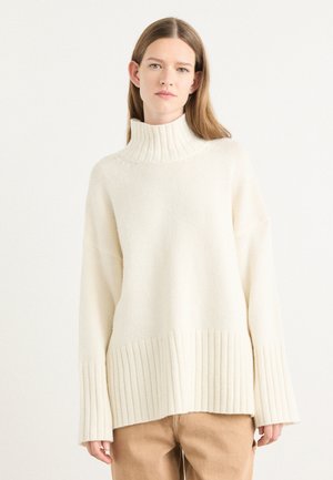 Pull en crème épais avec un col à côtes haut, des épaules tombantes et un ourlet à côtes. Texture en laine douce avec une coupe décontractée.