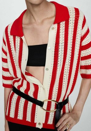 Osoba ubrana w czerwono-biały prążkowany sweter na guziki, pod którym znajduje się czarny krótki top, z czarnym paskiem z dużą złotą owalną klamrą.