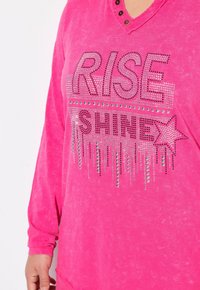 Top rosa a maniche lunghe con scollatura a V, caratterizzato da un testo in strass argentati "RISE SHINE" e un accento a forma di stella. Tessuto morbido con una texture invecchiata.