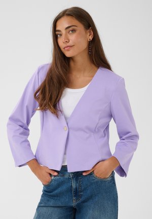 Femme aux longs cheveux bruns portant un blazer violet clair sur un haut blanc et un jean bleu, debout les mains dans les poches devant un fond uni.