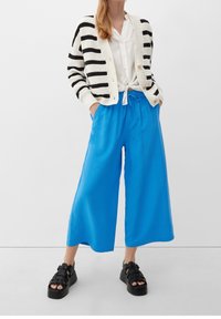 Gestreepte zwart-witte gebreide cardigan, witte blouse gestrikt in de taille, felblauwe wijd uitlopende broek en zwarte platformsandalen.