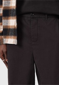 Pantaloni neri con una trama liscia, dotati di tasca frontale e chiusura con bottone superiore. Una camicia a quadri con tonalità marroni e nere è parzialmente visibile.