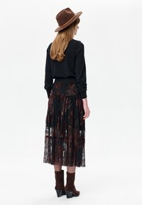 Vrouw met lang haar draagt een bruine hoed, een zwarte top met lange mouwen, een doorschijnende midi-rok met bloemmotief en bruine enkellaarsjes met hak, staand met de rug naar de kijker.