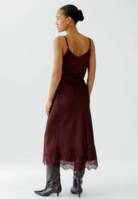 Robe en satin bordeaux avec un dos en V, bordée de dentelle au niveau de l'ourlet et des bretelles fines. Assortie à des bottes vernies jusqu'aux genoux. Texture lisse dans l'ensemble.