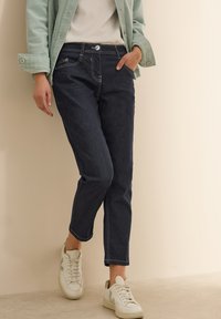 Jean en denim foncé avec des coutures claires, coupe slim, poches avant et fermeture à bouton. Portés avec des baskets blanches et une veste verte clair.