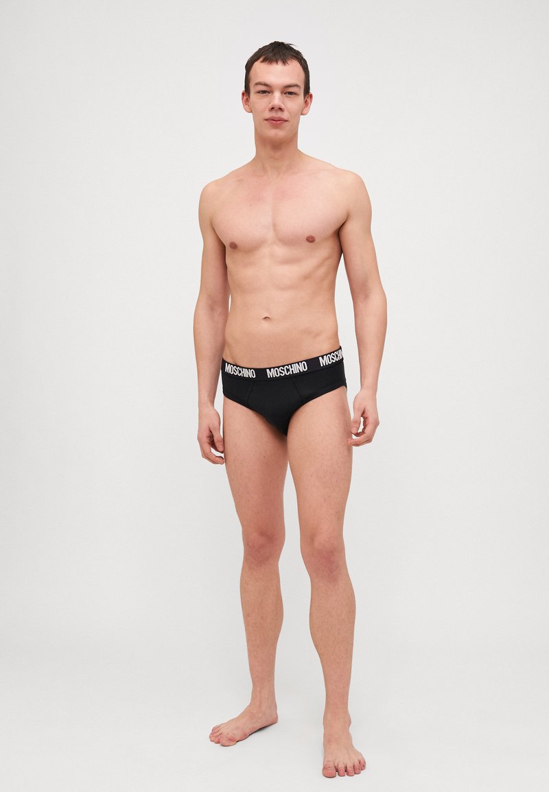 Boxer-briefs noirs pour hommes avec le logo Moschino sur la taille. Le tissu semble lisse et ajusté. Le modèle se tient pieds nus contre un fond clair.