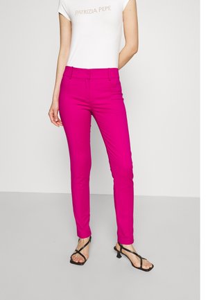 Vrouw draagt felroze aansluitende broek, wit T-shirt met korte mouwen en tekst, en zwarte sandalen met hak en bandjes, staande tegen een effen muur.