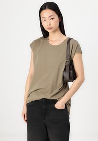 Tricou scurt din material moale, de culoare verde măslin, asortat cu blugi negri închis. O mică geantă de umăr maro are o curea și detalii cu buzunare.