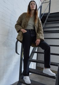 Chaqueta acolchada oliva sobre una blusa verde claro, combinada con jeans oscuros de cintura alta y zapatillas blancas, de pie en una escalera de metal.