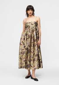 Γυναίκα με φόρεμα μέσης σε ελιά πράσινο, αμάνικο, με καφέ και μωβ λουλουδάτο σχέδιο, συνοδευόμενο από μαύρα επίπεδα παπούτσια, που στέκεται μπροστά σε λευκό φόντο.