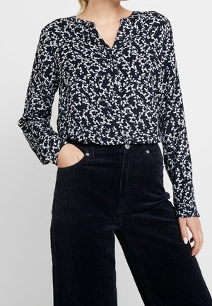 Femme portant un chemisier bleu marine à motif floral blanc et un pantalon en velours côtelé noir taille haute, une main dans la poche, sur un fond uni.