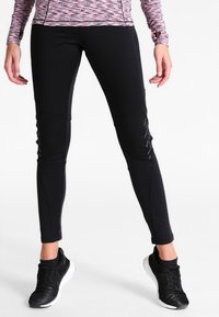 Schwarze Sportleggings mit schmalem Schnitt, glatter Textur und reflektierenden Akzenten an den Seiten. Kombiniert mit schwarzen Sneakers.