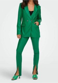 Completo verde aderente con giacca doppiopetto, pantaloni abbinati con spacchi sui orli e scarpe alte verde con cinturini. Texture liscia.