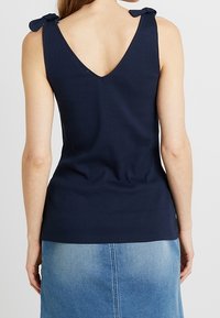 Marineblaues geripptes Tanktop mit V-Rücken und Schleifenakzenten an den Trägern, kombiniert mit einem hellen Jeansrock, der eine figurbetonte Silhouette zeigt.