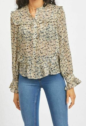 Kvinde iført en langærmet bluse med blomsterprint og flæsemanchetter og -kant, kombineret med tætsiddende blå jeans, står foran en ensfarvet hvid baggrund.