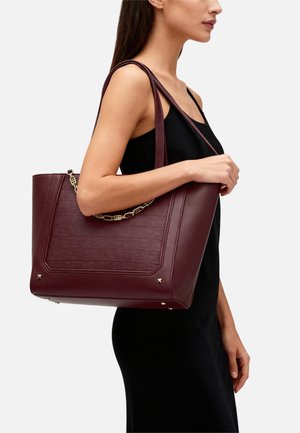 Donna con un abito nero che porta una grande borsa shopper in pelle bordeaux con dettagli a catena dorata sulla spalla.