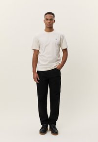 T-shirt en coton beige à manches courtes, avec un logo noir discret. Porté avec un pantalon noir et des chaussures noires, debout devant un fond neutre.