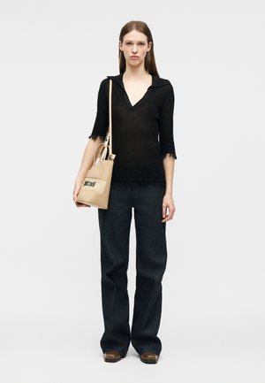 Vrouw die een zwart V-hals shirt met korte mouwen en een brede broek draagt, een beige handtas met schouderriem vasthoudend, staand tegen een witte achtergrond.