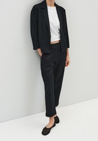 Femme portant un blazer noir, un pantalon noir, un top court blanc et des ballerines noires, debout contre un mur gris clair.