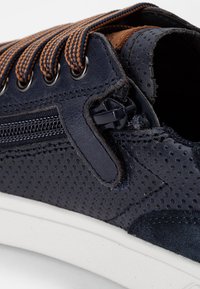 Zapatillas navy con diseño perforado, Acabados en color marrón claro, cordones naranjas en contraste y un detalle de cremallera en el lateral. Suela de goma blanca.