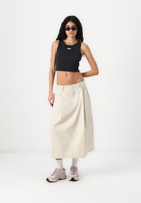 Black cropped tank top with white logo, beige flowing maxi skirt, and gray sneakers. Model wearing dark sunglasses and white socks.