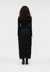 Μαύρο τοπ με μακριά μανίκια συνδυασμένο με μαύρη μάξι φούστα με φούντες, κατασκευασμένη από στρωμένο υφασμάτινο ύφασμα με υφή, φορεμένο με μαύρες μπότες μέχρι το γόνατο.