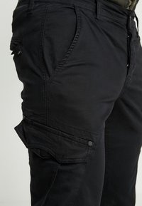 Närbild av svarta cargobyxor som visar sidfickan med klaff och knapp, samt detaljer i sömmarna på den framkrökta fickan.