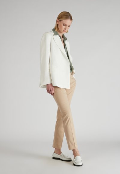 Witte blazer over een groene blouse, beige broek en witte instappers met zwarte zolen. Soepele texturen en een op maat gemaakte pasvorm.
