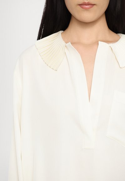 Cremefarbene Bluse mit einem gefältelten Kragen, V-Ausschnitt und einer einzelnen Brusttasche. Glatte Textur, lässige Passform und lange Ärmel.