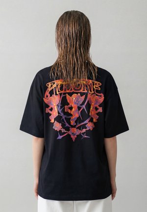 Person med vått hår som har på seg en svart T-skjorte med tre oransje keruber, et blomsteraktig torne-design og stilisert tekst på baksiden.