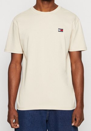 Beżowy bawełniany t-shirt z okrągłym dekoltem i krótkimi rękawami. Na lewej piersi znajduje się mała naszywka z logo w kolorach granatowym, czerwonym i białym.