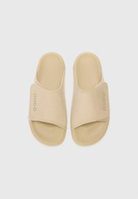 Beige glidesandaler med bred stropp som har "HUGO"-logoen. Laget av glatt materiale med flat såle og avrundede kanter.
