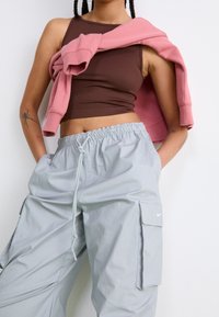 Person wearing a brown crop top, grey cargo pants with drawstring, and a pink sweatshirt draped over shoulders against a plain background.