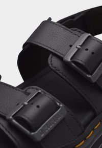 Μαύρο δερμάτινο σανδάλι με δύο ρυθμιζόμενες μεταλλικές αγκράφες και ορατή κίτρινη ραφή στην άκρη της σόλας.