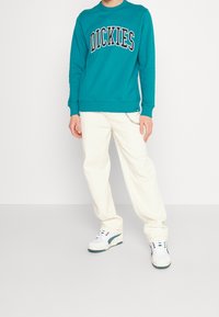 Sweatshirt teal avec le logo "DICKIES", associé à un pantalon texturé blanc et des baskets blanches avec des accents verts et un détail chaîne.