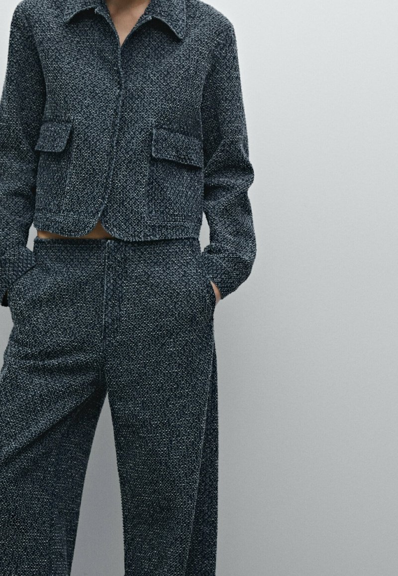 Giacca corta in tweed grigio scuro con due tasche frontali e colletto, abbinata a pantaloni a gamba larga coordinati, entrambi con motivo a trama.