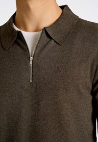 Brun zip-up tröja med klassisk krage, tillverkad av textilerad tyg. Har en subtil broderad logotyp och ett vitt inre lager.